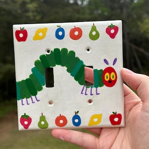 Peut inclure: Une plaque d'interrupteur double blanche ornée d'une chenille verte peinte à la main avec une tête rouge et des antennes violettes. Le motif comprend des illustrations colorées de divers fruits comme des pommes, des poires, des fraises et des citrons.