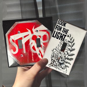 May include: Two light switch covers with different designs. One is a red and white stop sign with the text "STOP" and the other is black and white with the text "LOOK FOR THE LIGHT" and a butterfly design.