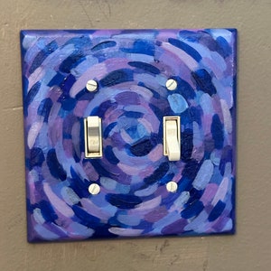May include: A square light switch cover hand-painted with a swirling pattern of blues and purples. The cover has two toggle switches and is mounted on a gray wall. The painting style features visible brushstrokes.