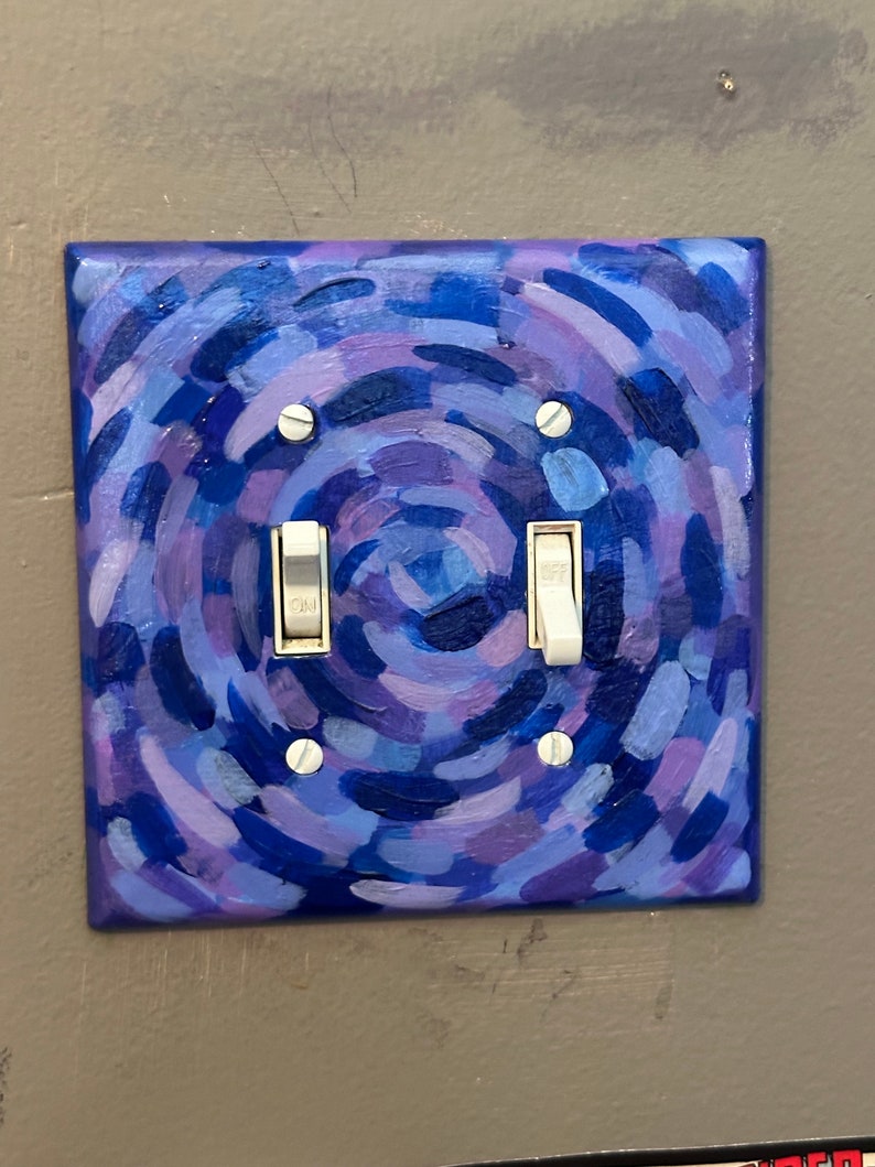 May include: A square light switch cover hand-painted with a swirling pattern of blues and purples. The cover has two toggle switches and is mounted on a gray wall. The painting style features visible brushstrokes.