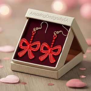 Puede incluir: Pendientes rojos con forma de lazo y ganchos dorados, presentados en una caja beige con el texto "AmazingDesignsBySA". Los pendientes se muestran sobre un fondo burdeos, con confeti rosa en forma de corazón esparcido.