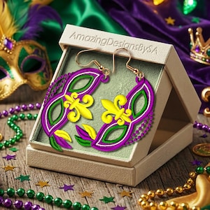 Könnte beinhalten: Ein Paar Ohrringe im Mardi Gras-Stil in einer Geschenkbox. Die Ohrringe sind maskenförmig und mit lila, grüner und gelber Stickerei versehen. Die Box ist von Perlen, einer Maske und einer Krone in festlichen Farben umgeben.