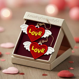 May include: A pair of heart-shaped earrings with the word "LOVE" embroidered in yellow on a red background. Each earring has white wings and gold-colored hooks. The earrings are displayed in a beige box with a maroon interior, surrounded by heart-shaped confetti.