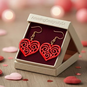 May include: Red heart-shaped earrings with a swirl pattern, displayed in a beige gift box. The earrings have gold-coloured hooks. The box is labelled "AmazingDesignsBySA". Decorative hearts and confetti are scattered on the wooden surface.