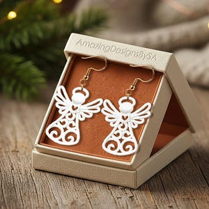 May include: White angel-shaped earrings with gold-colored hooks displayed in a beige box. The earrings have a detailed, openwork design. The box is open, revealing a brown interior. The background includes a wooden surface and a blurred Christmas tree.