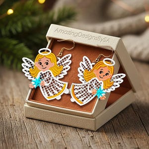 Puede incluir: Pendientes con forma de ángel con ganchos dorados, cabello rubio, alas blancas y una estrella azul. Los pendientes se muestran en una caja beige con el texto "AmazingDesignsBySA". Sobre una superficie de madera.