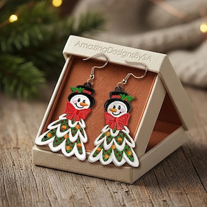 May include: A pair of snowman-shaped earrings displayed in a beige box. Each earring features a snowman face with a black hat, red bow tie, and a green Christmas tree-shaped body. The earrings are on a wooden surface.