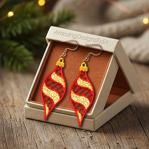 May include: A pair of Christmas ornament-shaped earrings in a display box. The earrings feature a red and gold design with a gold hook. The box is open, revealing the earrings against a brown interior. The text "AmazingDesignsBySA" is printed on the box.