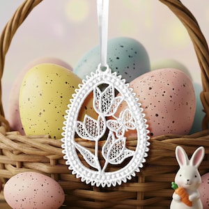 以下が含まれることがあります： 白いレースのイースターエッグオーナメント。花柄のデザインで、まだらのパステルカラーの卵が入った柳のバスケットの前に吊り下げられています。小さな白いウサギの置物がニンジンを持っています。