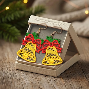 May include: A pair of Christmas bell earrings in a gift box. The earrings are yellow with red bows and green holly accents. The box is beige and the earrings are displayed on a wooden surface. The text "AmazingDesignsbySA" is on the box.