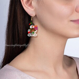 May include: A pair of Christmas-themed earrings featuring a snowman design. The snowman has a black hat, orange scarf, and holds a purple gift box. The earrings are white with gold-colored hooks. The text "amazingDesignBySA" is visible.
