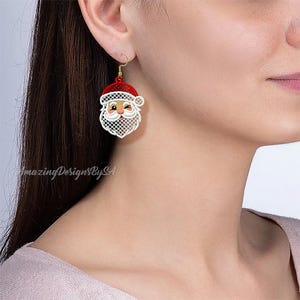 May include: A pair of Christmas-themed earrings featuring a Santa Claus design. The earrings have a red hat, a white beard, and a gold-colored hook. The Santa face is detailed with a winking eye and a smiling expression.