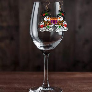 May include: A pair of snowman earrings with gold-colored hooks. Each earring features a snowman with a black top hat, a carrot nose, and a red and green scarf. The snowman is holding a purple gift box. The earrings are displayed in a wine glass.