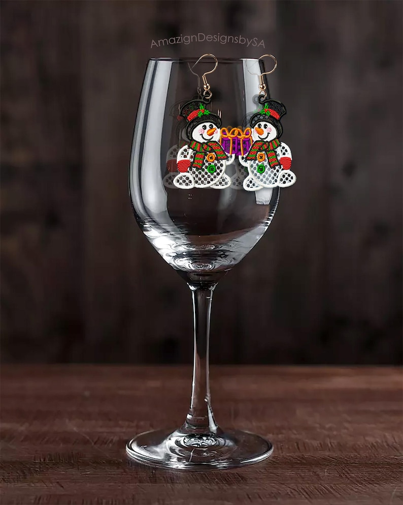 May include: A pair of snowman earrings with gold-colored hooks. Each earring features a snowman with a black top hat, a carrot nose, and a red and green scarf. The snowman is holding a purple gift box. The earrings are displayed in a wine glass.