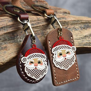May include: Two leather keychains with Santa Claus designs. One is teardrop-shaped, the other rectangular. Both have a red hat, white beard, and a winking face. The keychains are brown with metal hardware and stitched edges.