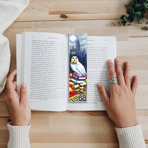 May include: A white owl with a letter in its beak sits on top of a stack of books. The owl is perched on a red and gold bookmark with a Hogwarts castle silhouette in the background. The bookmark is made of cross-stitch.