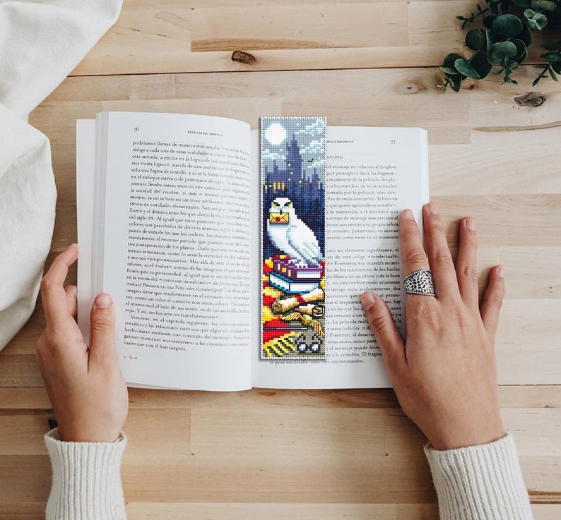 May include: A white owl with a letter in its beak sits on top of a stack of books. The owl is perched on a red and gold bookmark with a Hogwarts castle silhouette in the background. The bookmark is made of cross-stitch.