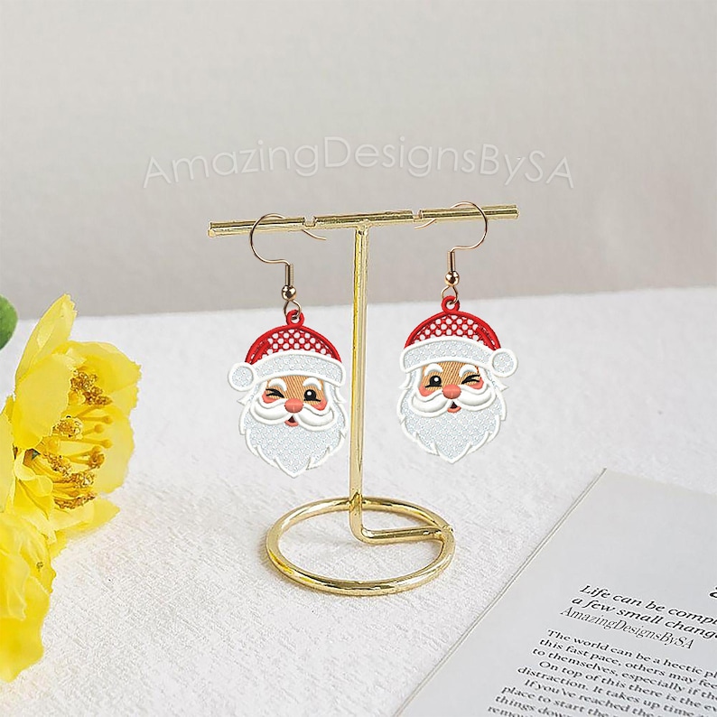 May include: A pair of Christmas-themed dangle earrings featuring Santa Claus faces. Each earring has a red hat with white trim, a white beard, and a smiling face. The earrings are displayed on a gold-colored stand.