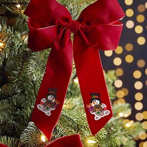 May include: A large, festive red velvet bow with long, flowing tails, adorned with embroidered snowman appliques. The bow is displayed on a Christmas tree, creating a holiday decoration.