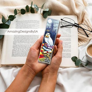 May include: A white owl with yellow eyes sits on top of a stack of books. The owl is holding a letter in its beak. The bookmark is a colorful illustration with a night sky background and a castle in the distance. The bookmark is made of fabric and has a white border.
