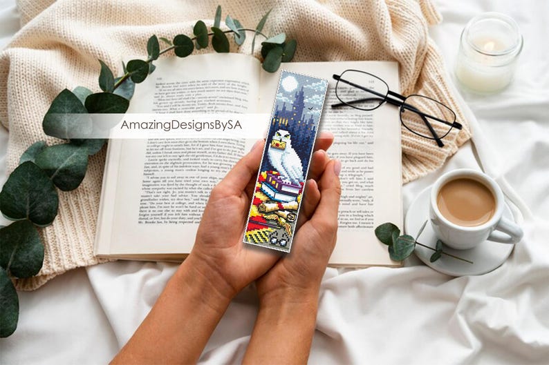 May include: A white owl with yellow eyes sits on top of a stack of books. The owl is holding a letter in its beak. The bookmark is a colorful illustration with a night sky background and a castle in the distance. The bookmark is made of fabric and has a white border.