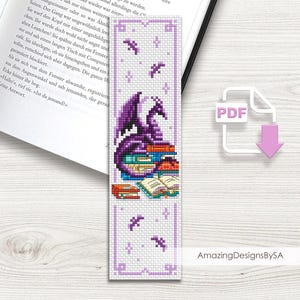 May include: A cross-stitch bookmark with a purple dragon design sitting on books. The bookmark has a light purple border with decorative elements and the text "AmazingDesignsBySA". The design includes stars and small dragon silhouettes.