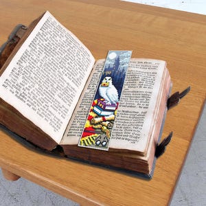 May include: A bookmark featuring a white owl perched on a stack of books with a Hogwarts castle background. The bookmark is made of fabric and has a colorful design.