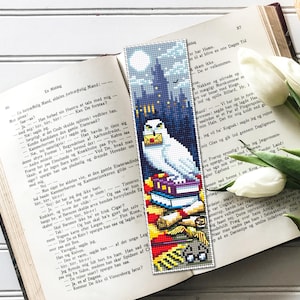 May include: A white owl bookmark with a stack of books, scrolls, and a pair of glasses. The owl is perched on a stack of books with a red and gold scarf. The background is a dark blue and purple scene with a castle in the distance.