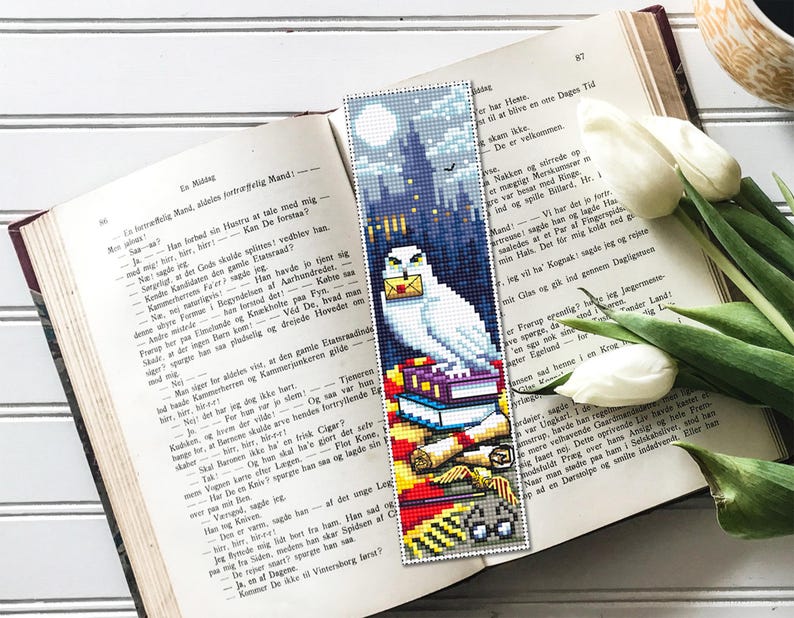 May include: A white owl bookmark with a stack of books, scrolls, and a pair of glasses. The owl is perched on a stack of books with a red and gold scarf. The background is a dark blue and purple scene with a castle in the distance.