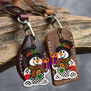 May include: Two leather keychains with a snowman design. The snowmen have black top hats, orange carrot noses, and red mittens. One keychain is teardrop-shaped, the other is rectangular. Both have a metal clasp and a brown leather strap.