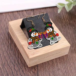 May include: A pair of snowman earrings with gold-colored hooks. Each earring features a smiling snowman wearing a black top hat, a red and green scarf, and holding a purple gift. The earrings are displayed on a black card inside a wooden box.
