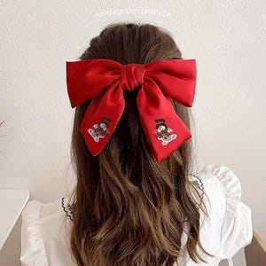 May include: A large, vibrant red bow hair clip. The bow features two embroidered snowman designs on the tails. The hair accessory is worn in the hair, with the back of the head and shoulders visible. The bow is made of a silky material.