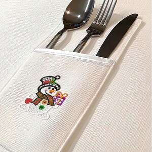 May include: A white cloth cutlery holder with a snowman design. The snowman has a black top hat, a red and green scarf, and is holding a purple gift. The holder contains a spoon, fork, and knife.