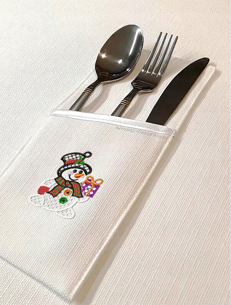 May include: A white cloth cutlery holder with a snowman design. The snowman has a black top hat, a red and green scarf, and is holding a purple gift. The holder contains a spoon, fork, and knife.