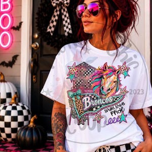Pode incluir: Uma t-shirt branca de manga curta com uma estampa gráfica colorida de uma cabeça de cavalo, uma torre de água e estrelas. O design inclui o texto "Broncos" e "small town pride" numa mistura de letras cursivas e de forma com detalhes de leopardo.