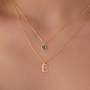 May include: Two gold necklaces layered together. The top necklace has a small heart pendant with a teal center. The bottom necklace has a gold letter "E" pendant. The necklaces are on a person's neck.
