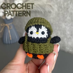 May include: A handmade crochet penguin wearing a green hat and sweater. The penguin has a white face with black and yellow details. The text "CROCHET PATTERN" is at the top of the image.