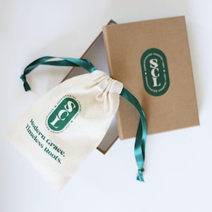 May include: A white drawstring bag with green text that reads "Modern Grace, Timeless Roots." The bag is tied with a green ribbon and is sitting on top of a brown box with a green logo that reads "SCL Sophisticated Country Lady".