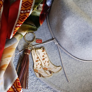 May include: A gold-tone keychain with a brown leather tassel and a pair of white cowboy boots with floral embroidery. The keychain is attached to a gray felt hat.