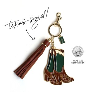 May include: A gold-tone keychain with a brown leather tassel and a pair of green and brown enamel cowboy boots. The boots are about the size of a US quarter, as shown in the image. The keychain has the text "texas-sized!" written on it.