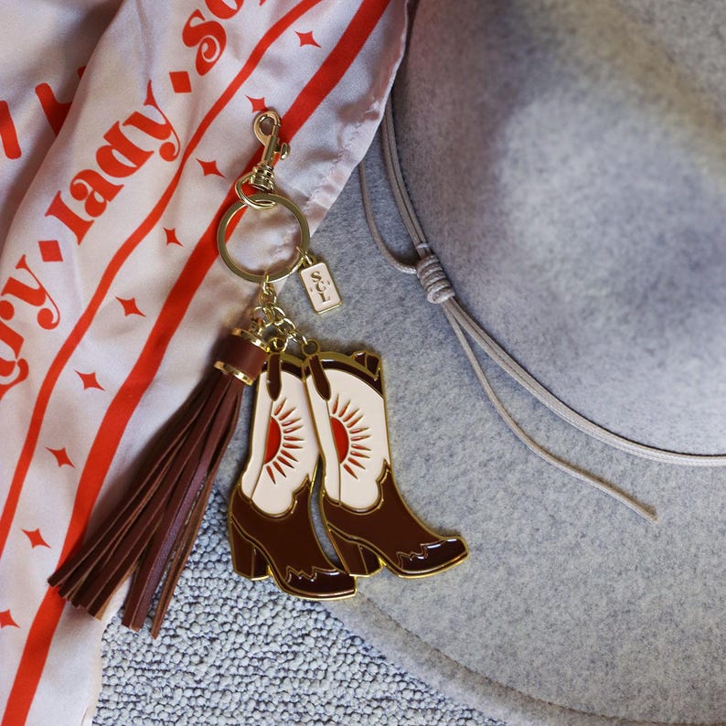 May include: A gold-tone keychain with a brown leather tassel and two enamel cowboy boot charms. The boots are white with a red sun design and brown heels.