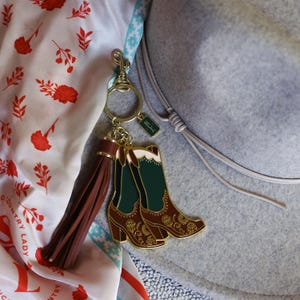 May include: A gold-tone keychain with a brown leather tassel and a charm of two green and brown cowboy boots. The boots have gold embroidery and a white accent at the top.