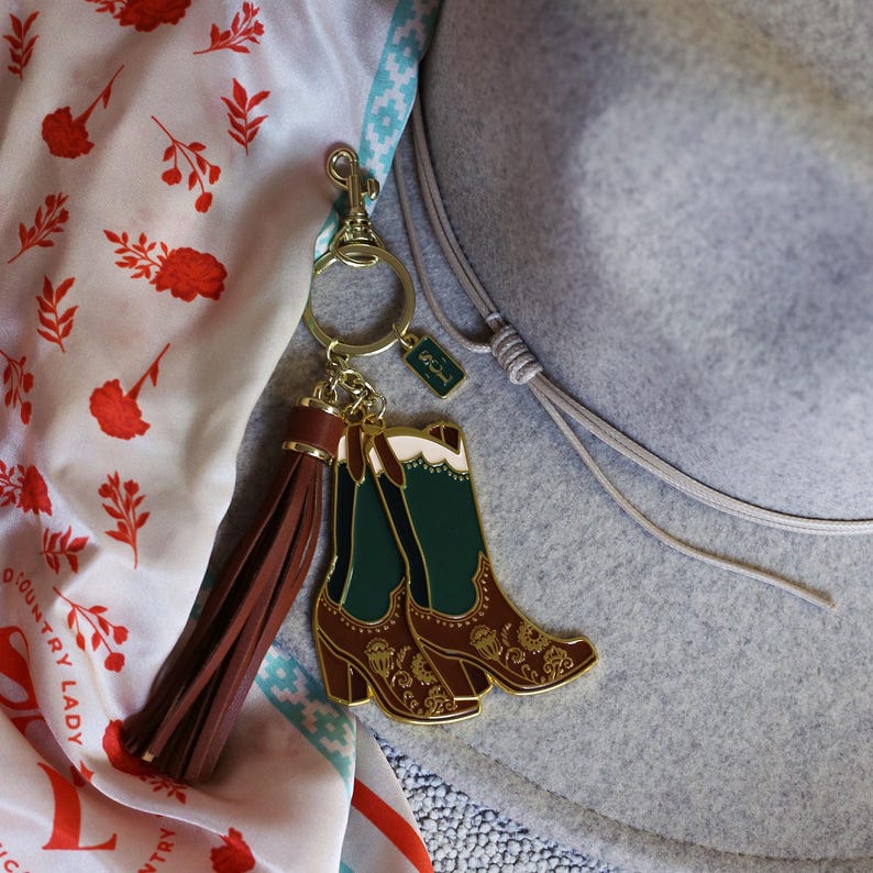 May include: A gold-tone keychain with a brown leather tassel and a charm of two green and brown cowboy boots. The boots have gold embroidery and a white accent at the top.