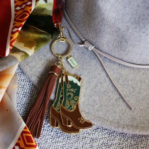 May include: A gold-tone keychain with a brown tassel and a charm of two green and brown cowboy boots with a paisley pattern. The charm is attached to a gold-tone ring and a small tag with the text "L21".