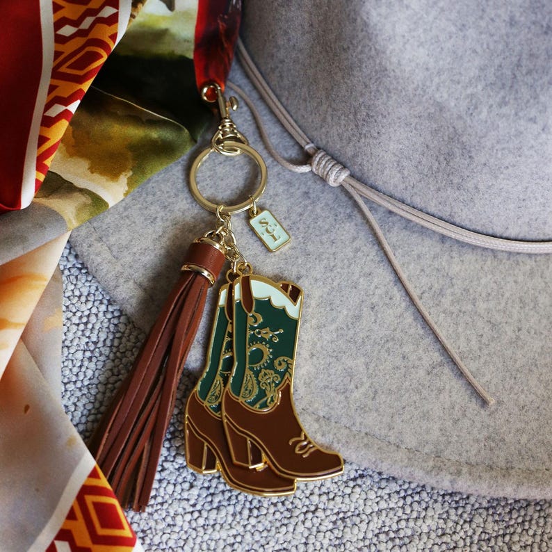 May include: A gold-tone keychain with a brown tassel and a charm of two green and brown cowboy boots with a paisley pattern. The charm is attached to a gold-tone ring and a small tag with the text "L21".