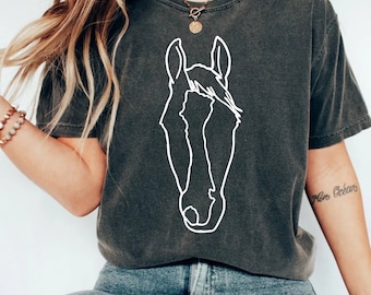 May include: A dark gray t-shirt with a white outline of a horse's head. The shirt is worn by a person with long brown hair and a gold necklace. The person is wearing blue jeans.