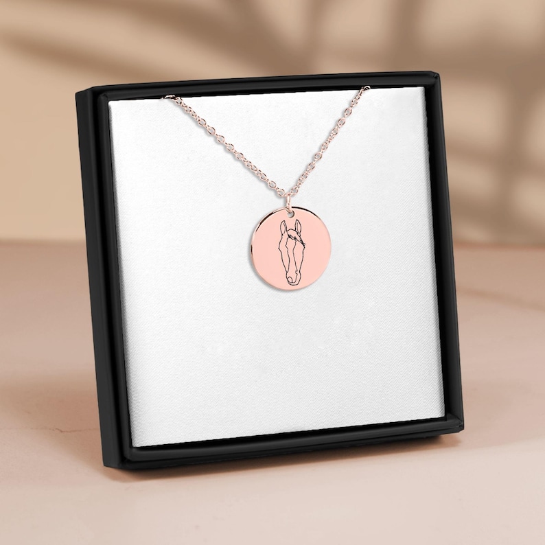 Puede incluir: Un collar de cadena de plata con un colgante redondo que presenta un dibujo lineal de la cabeza de un caballo. El colgante es de color oro rosa.