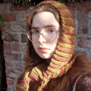 May include: A hooded scarf featuring a ribbed knit texture with alternating horizontal stripes in olive green and burnt orange. The garment covers the head and wraps around the neck, showcasing a consistent yarn pattern throughout the entire piece.