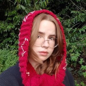 May include: A vibrant red crochet hood with a ribbed texture and white stitched accents along the edges. The hood features long, integrated scarf-like ends that drape downwards, crafted from thick yarn with a visible, chunky knit pattern throughout.