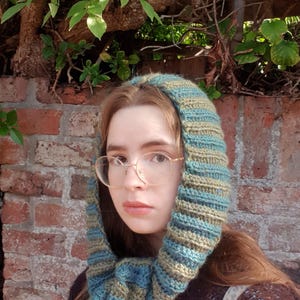 May include: A crochet hood scarf featuring horizontal stripes in alternating shades of teal, blue, and olive green. The accessory is constructed with a ribbed texture and long ends that drape down, providing full coverage for the head and neck area.